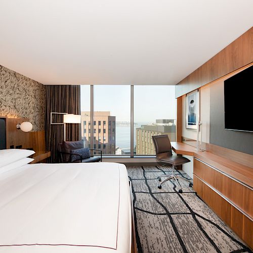 A modern hotel room with a large bed, a desk with a chair, a flat-screen TV, and a window offering a city view.