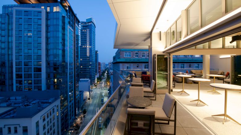 A modern cityscape at dusk with tall buildings and a well-furnished balcony featuring tables and chairs, overlooking a street below.