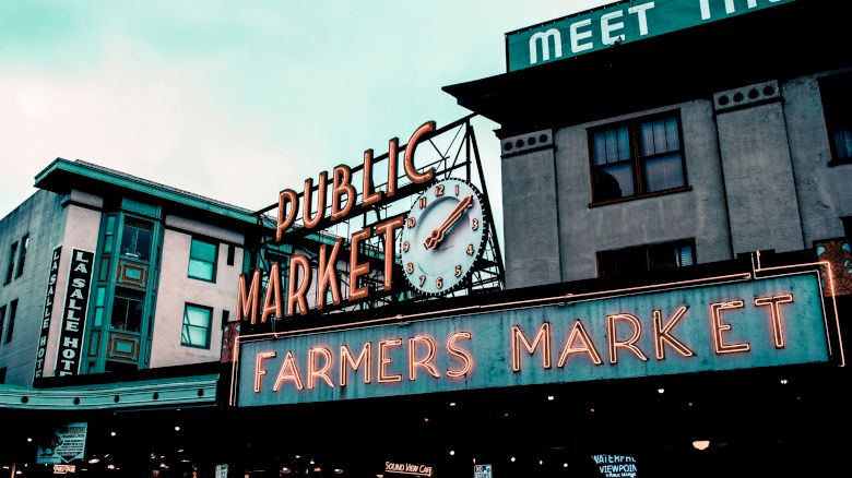 The image shows the iconic neon signs for "Public Market" and "Farmers Market" at Pike Place Market in Seattle, Washington, with adjacent buildings.