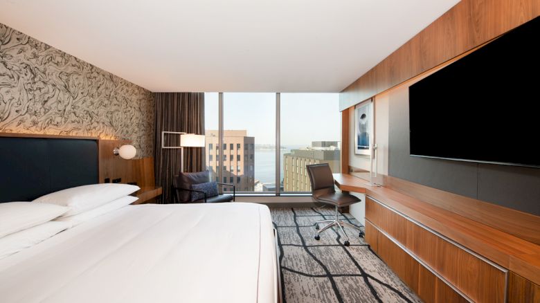 A modern hotel room with a large bed, a wall-mounted TV, a work desk with a chair, and a window view of buildings and a body of water.