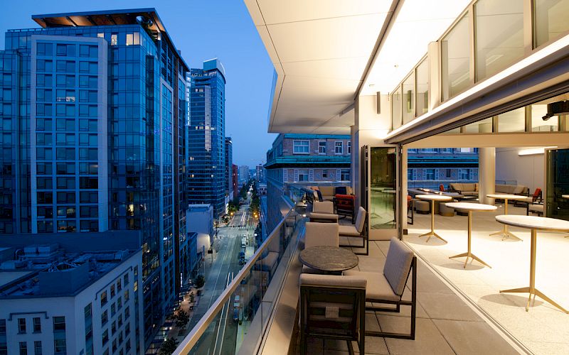 A modern rooftop terrace overlooking a cityscape with tall buildings, featuring outdoor seating and a well-lit indoor area.