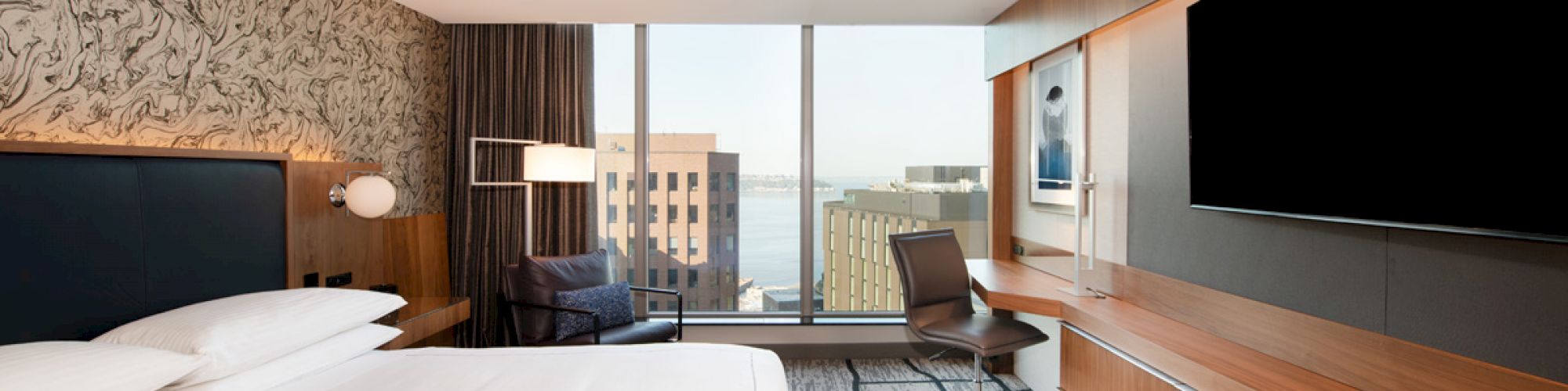 A modern hotel room features a king-sized bed, a large flat-screen TV, a desk with a chair, and a panoramic window with a city view.