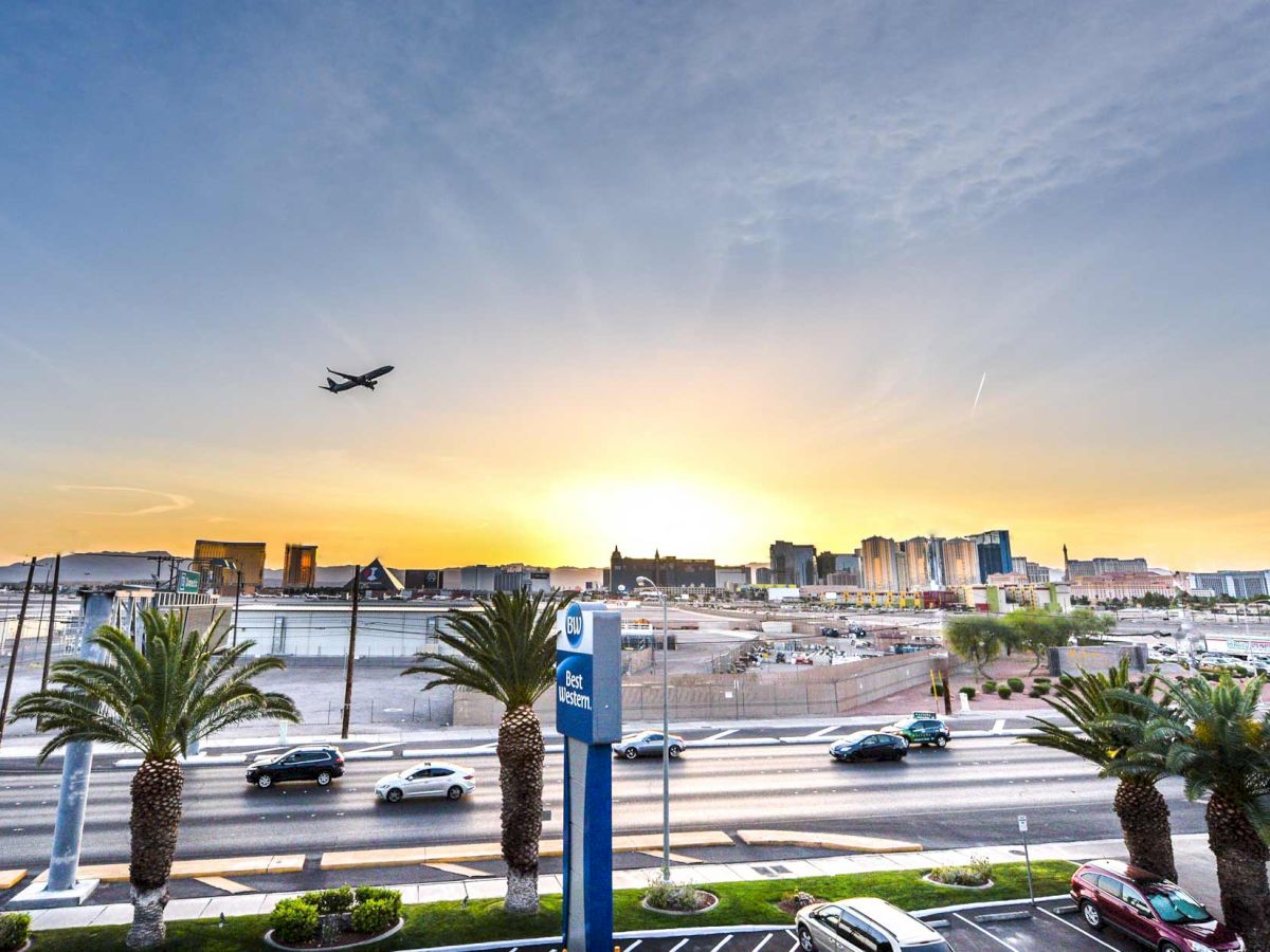 A sunset view of a cityscape, with an airplane above, palm trees, a hotel sign, a busy street, parked cars, and distant Ferris wheel.