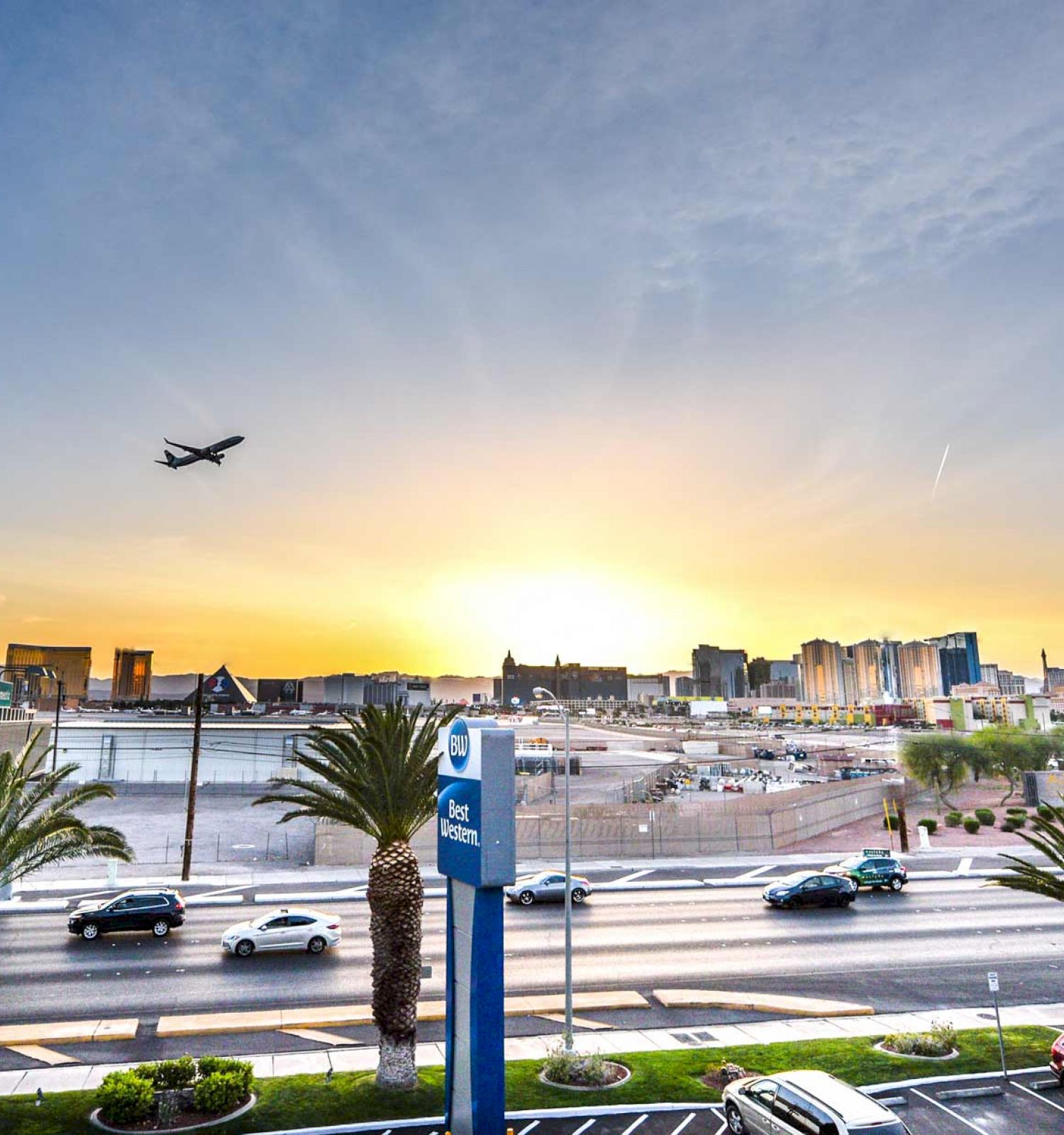 A sunset view of a cityscape, with an airplane above, palm trees, a hotel sign, a busy street, parked cars, and distant Ferris wheel.