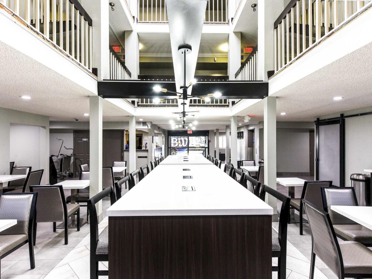 The image shows a modern indoor dining area with tables and chairs, and a long center table. The space features high ceilings and a clean design.