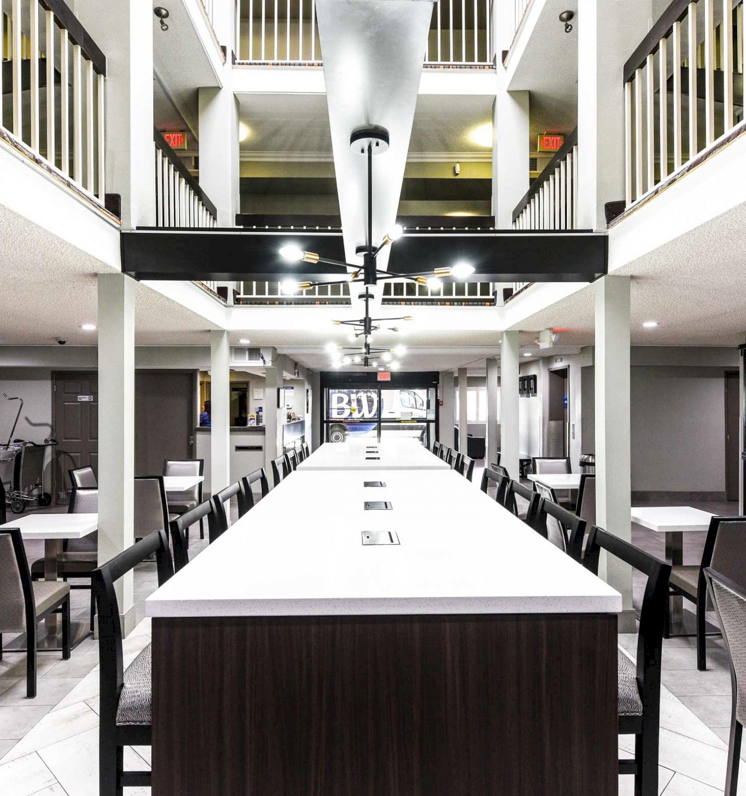 The image shows a modern indoor dining area with tables and chairs, and a long center table. The space features high ceilings and a clean design.