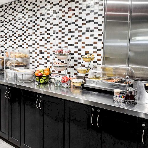 The image shows a breakfast buffet area with a variety of food and beverage options, including pastries, fruits, and a waffle maker.