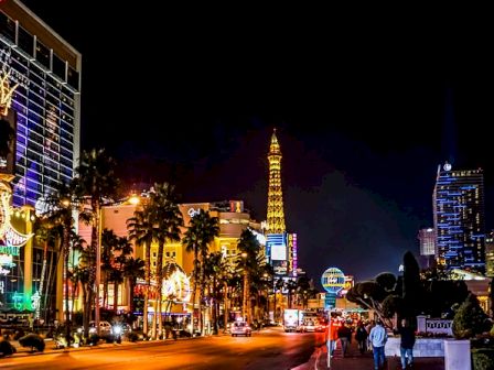 A vibrant and illuminated cityscape at night featuring high-rise buildings, palm trees, and a replica of the Eiffel Tower in the background.