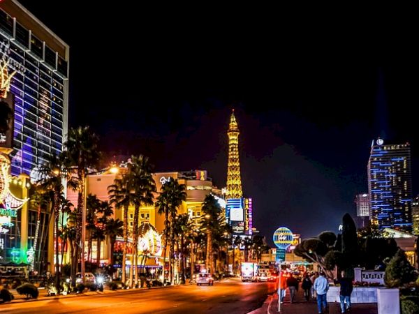 A vibrant and illuminated cityscape at night featuring high-rise buildings, palm trees, and a replica of the Eiffel Tower in the background.
