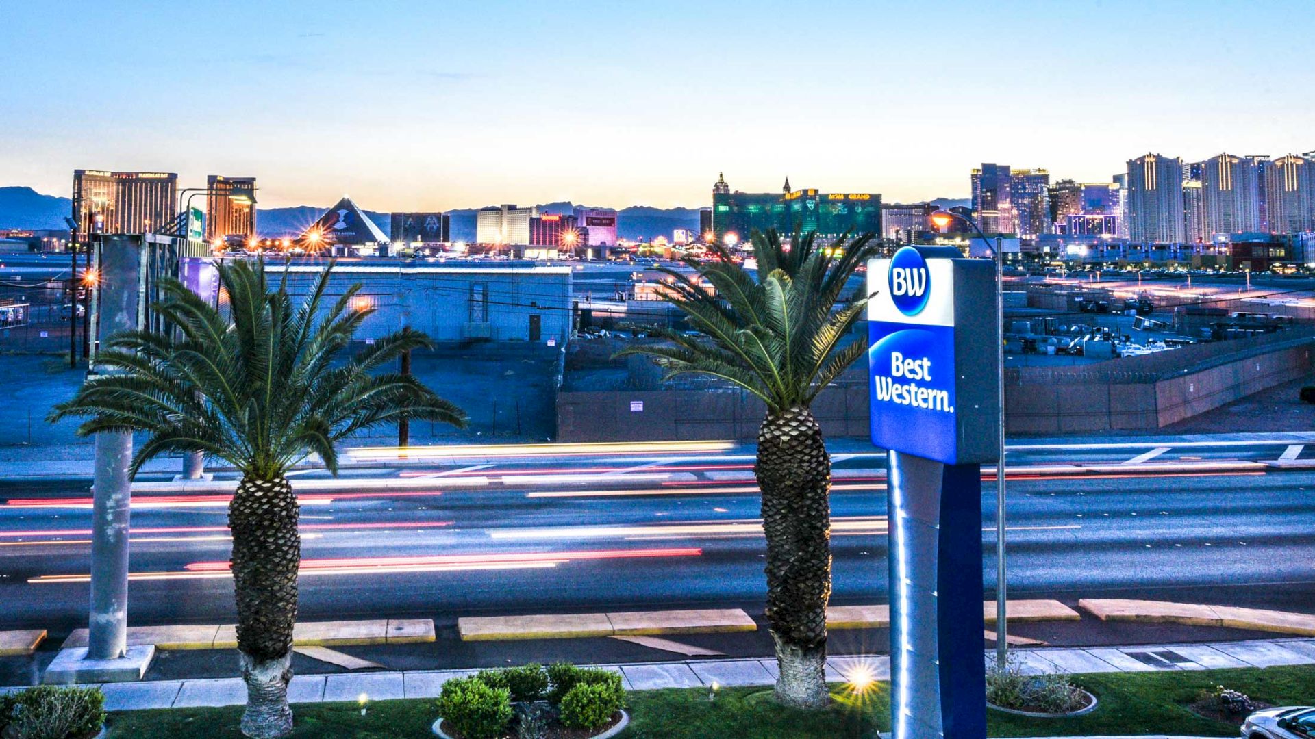 The image shows a street with palm trees and a Best Western hotel sign in the foreground, and a city's skyline with various buildings in the background.