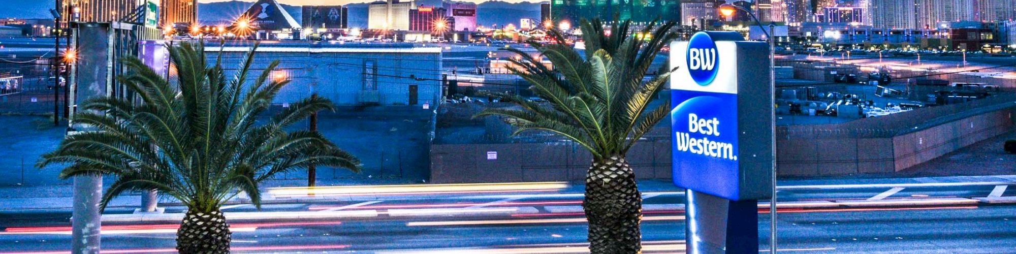 The image shows a street with palm trees and a Best Western hotel sign in the foreground, and a city's skyline with various buildings in the background.